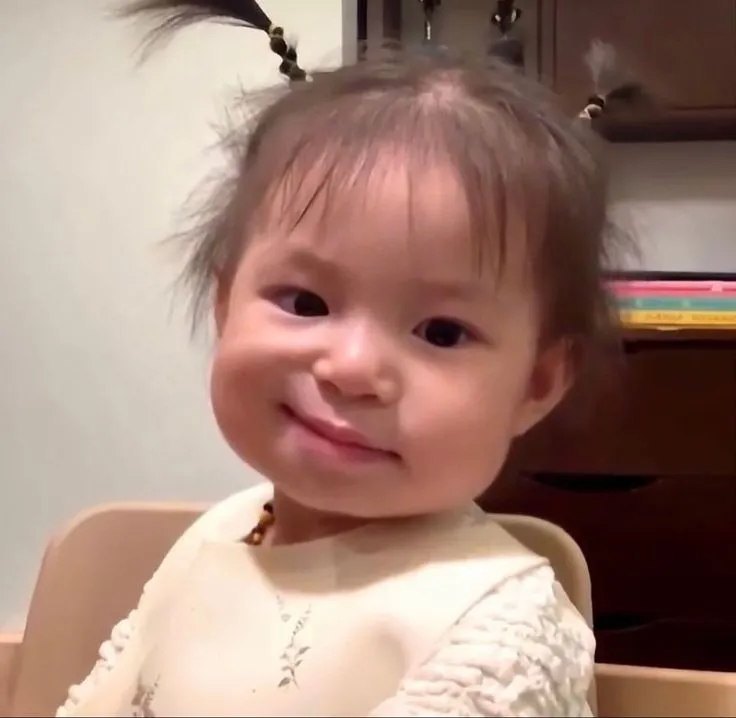 25 Cô bé mặc váy trắng ngồi trong nhà cười mím môi, ánh mắt tinh nghịch như vừa nghe chuyện vô lý mà vẫn lịch sự