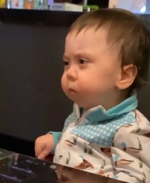 7 Cậu bé mặc áo thun họa tiết đeo yếm xanh ngồi đơ mặt nhìn sang ngang với vẻ chịu đựng và cạn lời trước tình huống oái oăm.