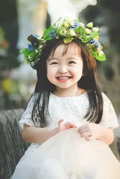 6 Trong váy trắng và vòng hoa xanh, bé gái cười rạng rỡ giữa nền cây mờ, dịu dàng như nàng thơ nhí